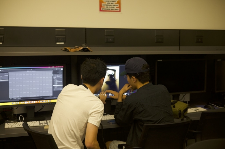 Two Tisch Summer High School Photography students sit in a computer lab pointing at a sepia-toned image that is being edited on a computer screen.